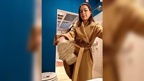 Media: Video of a woman in a tan trench coat, adjusting her hair in a modern bathroom with blue walls, white tiles, and a striped rug.