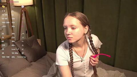 Media: A video of a young woman with long brown braids, wearing a white top, sitting on a bed with green curtains, holding a pink feather duster, with a digital camera overlay.