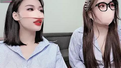 Media: Video of two young women with pale skin, short dark hair, wearing light blue striped shirts and face masks, seated on a gray couch in a plain room.