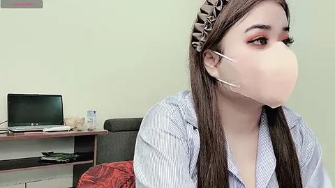 Media: Video of an Asian woman with long brown hair wearing a light blue striped shirt, beige face mask, and a headband, sitting indoors with a laptop and papers on a desk.