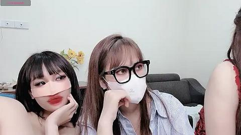 Media: Video of two Asian women with fair skin, one with red lipstick, wearing glasses and mask, and the other with straight hair, in a modern living room with white walls and a gray couch.