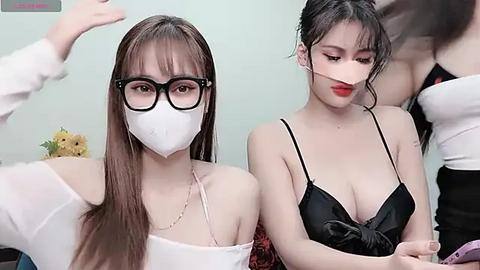 Media: Video of two East Asian women in casual attire, one wearing a mask and glasses, the other a black top, indoors with flowers in the background.