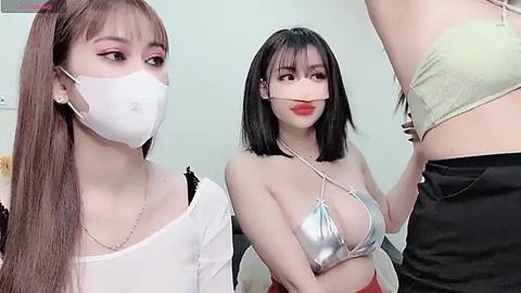 Media: Video of two East Asian women with light skin, one in a white mask, silver bikini top, and red skirt, the other in a green top, standing in a room with a pale wall.