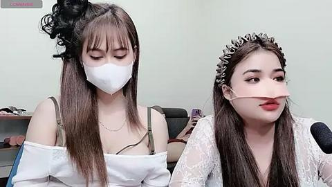 Media: Video of an Asian woman with long brown hair, wearing a white face mask, off-shoulder lace top, and a crown, seated indoors.