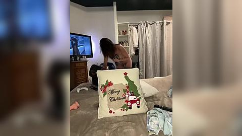 Media: Video of a woman with long hair in a living room, wearing a black top, bending over a beige pillow with \"Merry Christmas\" and holiday decorations.