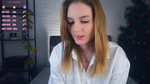 Media: Video of a young Caucasian woman with light skin, long blonde hair, and light makeup, wearing a white shirt, standing indoors near a Christmas tree and a white brick wall.