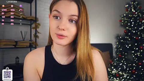 Media: Video of a young Caucasian woman with fair skin and long blonde hair, wearing a black tank top, standing in a modern living room with a decorated Christmas tree.