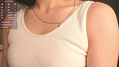 Media: Video of a light-skinned woman wearing a white ribbed tank top, showcasing a thin gold necklace, with a social media app interface in the background.