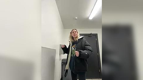 Media: Video of a blonde woman in a black puffer jacket, blue jeans, and a green scarf, walking down a brightly lit, modern hallway with white walls and a fluorescent light fixture.