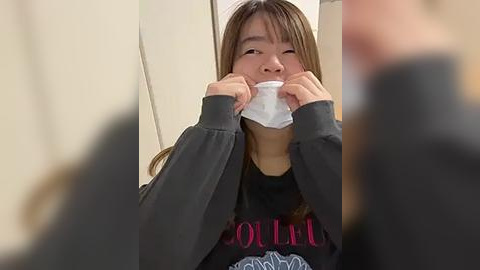 Media: Video of a young Asian woman in a gray sweater, wearing a white surgical mask, covering her nose and mouth with both hands, in a blurred indoor setting.