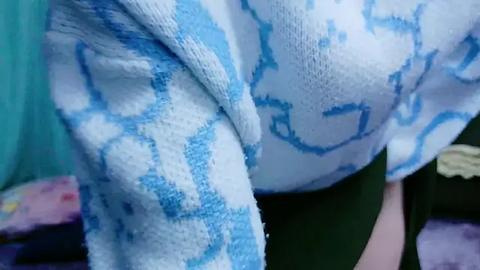 Media: Close-up video of a person wearing a white, blue patterned towel with a geometric design, partially covering their chest. The background includes colorful fabric, likely clothing or bedding, suggesting a casual, relaxed setting.