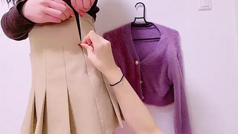 Media: Video of a person wearing a beige pleated skirt, adjusting a black belt, while another hand holds a purple cardigan with a black hanger in a white room.