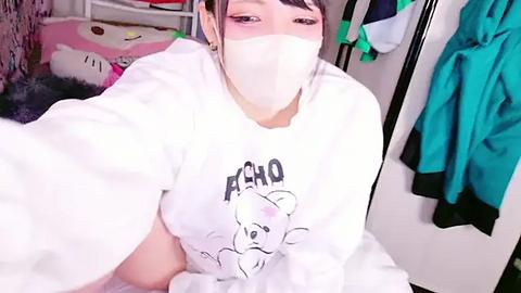 Media: Video of a woman with light skin and short, dark hair, wearing a white mask, sweatshirt with a bear graphic, and taking a selfie in a messy room.