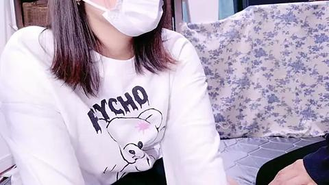 Media: A video of a person wearing a white face mask, white long-sleeve shirt with a cartoon bear graphic, and black pants, seated in a room with a blue floral-patterned couch and a wooden table in the background.