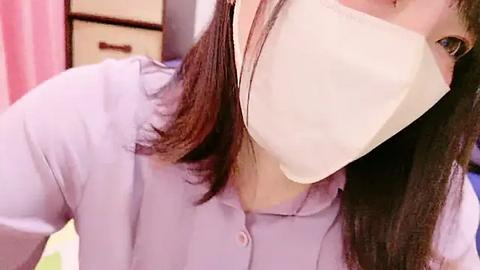 Media: A video of a person wearing a large, pale yellow face mask over their mouth and nose, with long, dark brown hair visible. They are dressed in a light purple, button-up shirt. The background includes a pink curtain and wooden furniture.