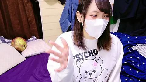 Media: Video of an East Asian woman with shoulder-length dark hair, wearing a white sweatshirt with a cartoon bear, a surgical mask, and a pair of white headphones, in a cluttered bedroom with disorganized clothes and a stuffed animal.