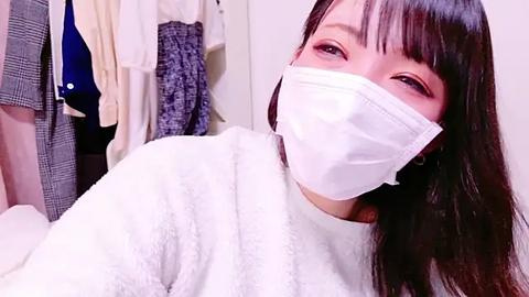 Media: Video of an East Asian woman with long dark hair, wearing a white surgical mask, white sweater, and a gray jacket in a cluttered closet.