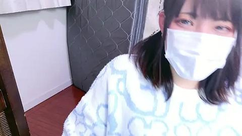 Media: Video of an Asian woman with straight, shoulder-length dark hair, wearing a white surgical mask, a blue-patterned hospital gown, and standing in a hospital room with a gray quilted blanket and white walls.