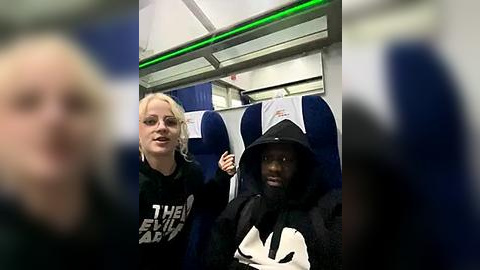 Media: Video of a woman with blonde hair and a man in a black hoodie and Adidas shirt, seated in an airplane.