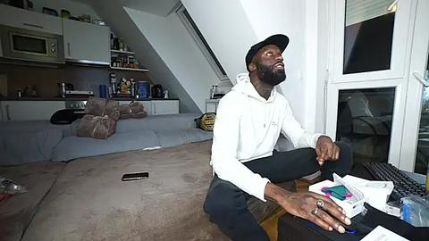 Media: Video of a seated man in a white hoodie, black pants, and cap, looking thoughtful in a modern, cluttered loft with exposed beams, kitchen, and a window.