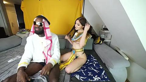 Media: Video of a black man in a white sweatshirt and pink headscarf, sitting on a gray couch, with a white woman in a gold bikini and headscarf braiding his hair.