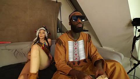 Media: Video of a dark-skinned African man in traditional attire, sitting beside a light-skinned woman in a tribal costume, in a dimly lit attic room with a beige curtain and black backdrop.