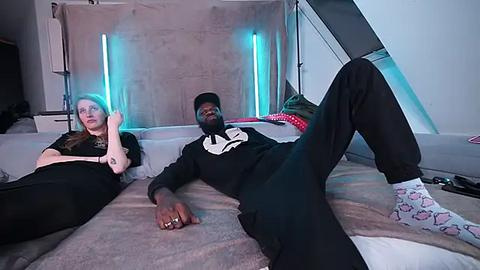 Media: Video of a tall, muscular Black man in a black hoodie and pants, lying on a beige sofa, with a blonde woman in a black top and white socks with pink hearts. Background features a modern, minimalist room with blue neon lights and a staircase.