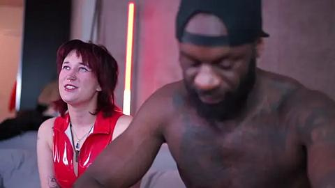 Media: Video of a pale, red-haired woman in a shiny red PVC dress, standing beside a shirtless, muscular black man with a backward cap, in a dimly lit room.