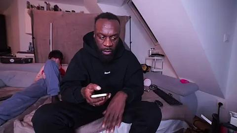 Media: Video of a black man in a black hoodie and jeans, sitting on a bed in a dimly lit attic room, focused on a phone.