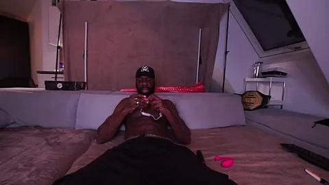 Media: Video of shirtless, muscular Black man in black hat and pants, playing video game on a bed in dimly lit room with beige walls, red light, and gaming controllers.