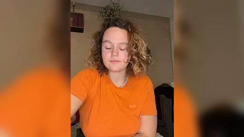 Media: Video of a young woman with curly, shoulder-length hair, wearing an orange t-shirt, sitting in a dimly lit room with beige walls and a plant hanging from the ceiling.