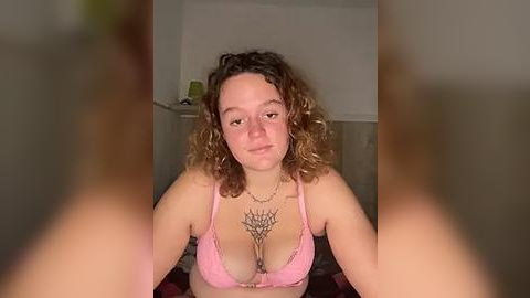 Media: A video of a young woman with curly brown hair and fair skin, wearing a pink bra, sitting indoors with a background of beige walls and a green cup.