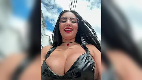 Media: Video of a confident, voluptuous woman with long black hair, wearing a shiny black latex dress with a plunging neckline, and clear glasses, standing under a blue sky with white clouds.