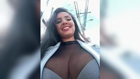 Media: Video of a smiling, curvaceous Black woman with long black hair, wearing glasses and a black choker, in a low-cut gray top, standing in a city street.