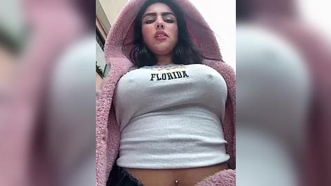 Media: Video of a curvy woman with large breasts, wearing a tight, white crop top with \"Florida\" printed on it and a pink fuzzy robe. She has a belly button piercing. Background is blurry.