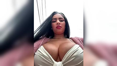 Media: Video of a curvaceous woman with long black hair, wearing a low-cut white top that accentuates her ample cleavage, a pink cardigan, and a pearl necklace.