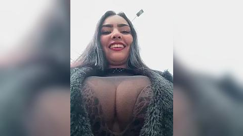 Media: A video of a smiling, light-skinned woman with long, straight black hair, wearing a sheer, lace top that reveals her large breasts. She is in a furry coat, and the background is blurred.