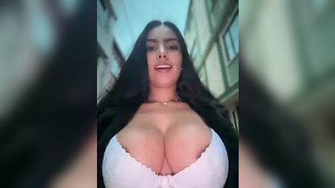 Media: Video of a smiling woman with long black hair, wearing a white bra that accentuates her large breasts, standing in a narrow, blurred alleyway with stone buildings in the background.