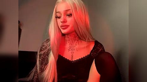 Media: Video of a young woman with long, platinum blonde hair, wearing a black lace top, adorned with intricate tattoos on her neck and chest. She has a pale complexion and is bathed in a warm, red-hued lighting.