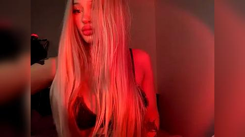 Media: Video of a woman with long, platinum blonde hair, wearing a black bra, under red lighting, in a dimly lit room. Her skin is fair, and she has full lips.