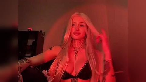 Media: Video of a blonde woman with long hair, wearing a black bra and necklace, sitting in a dimly lit room with red lighting.