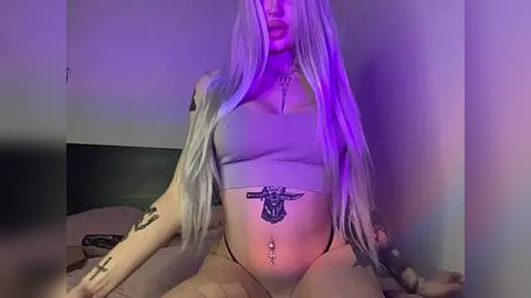 Media: Video of a pregnant woman with long, platinum blonde hair, wearing a light grey crop top, black thong, and multiple tattoos, sitting on a bed with a purple glow.