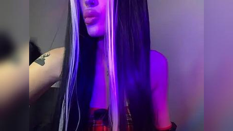 Media: Video of a young woman with long, straight, black hair streaked with white. She has a tattoo on her upper arm and wears a red plaid shirt. The background is a plain, neutral wall with purple and blue lighting.