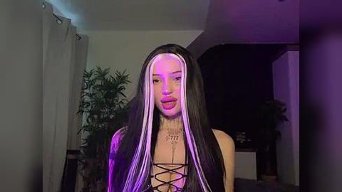 Media: A video of a young woman with long, straight black hair and white streaks, wearing a black corset top, standing in a dimly lit room with purple lighting, greenery, and white curtains.