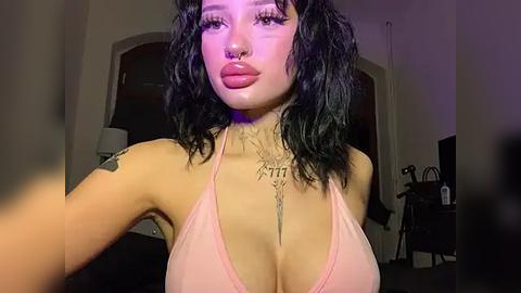 Media: Video of a young woman with fair skin, black curly hair, and a large nose ring. She wears a pink halter top, highlighting her ample cleavage. The background is a dimly lit room with a computer and a lamp.