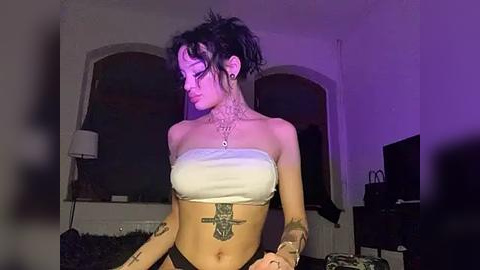 Media: Video of a pale-skinned woman with short dark hair, tattoos on her arms and chest, wearing a white strapless top, standing indoors under purple lighting.