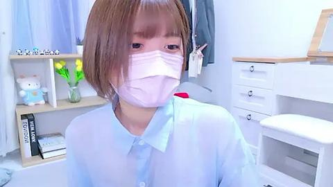 Media: A video of an Asian woman with short brown hair, wearing a light blue shirt, a white face mask, and seated in a brightly lit, minimalist bathroom with white cabinets, a plant, and a stuffed bear.