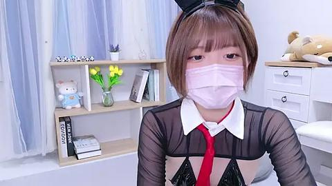 Media: Video of an Asian woman with short brown hair, wearing a black mesh top, red tie, white mask, black bunny ears, and white collar, in a light, cozy bedroom with white furniture, stuffed animals, and potted plants.