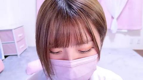 Media: Video of an Asian woman with straight, shoulder-length brown hair, wearing a light pink surgical mask, sitting in a pastel pink room with drawers and a white bed in the background.