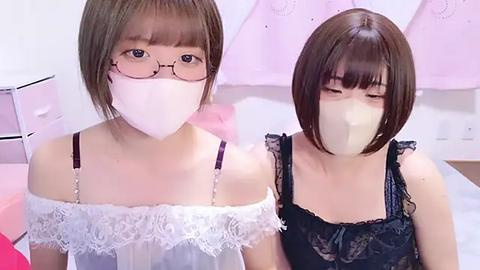 Media: Video of two young Asian women with short brown hair, wearing white face masks, glasses, and lace-trimmed camisoles, sitting close in a softly lit room with pastel decor.